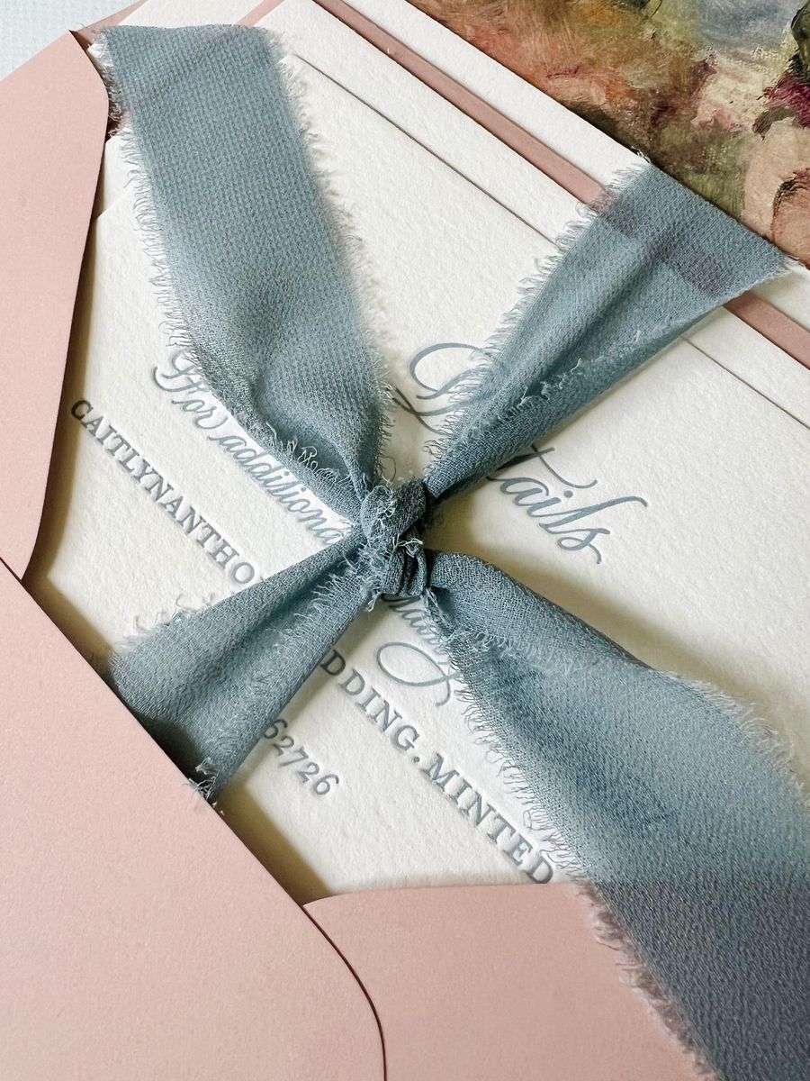 A silk ribbon tied around a letterpress suite resting in a blush envelope